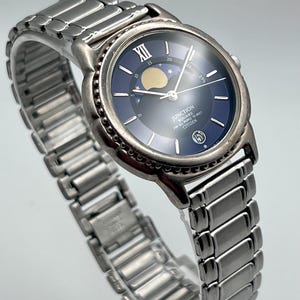 May include: A silver Citizen Junction Square watch with a blue face and a moon phase indicator. The watch has a stainless steel strap and a round face with Roman numerals at the 12 o'clock position. The watch face also displays the date.