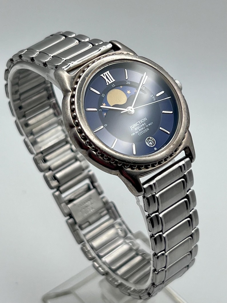 May include: A silver Citizen Junction Square watch with a blue face and a moon phase indicator. The watch has a stainless steel strap and a round face with Roman numerals at the 12 o'clock position. The watch face also displays the date.