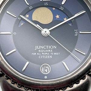 May include: Close-up of a Citizen Junction Square watch. The watch face is a gradient of dark blue to grey, featuring a moon phase indicator with a gold moon and stars. The watch has silver hands and markers, with the date displayed at the 6 o'clock position.