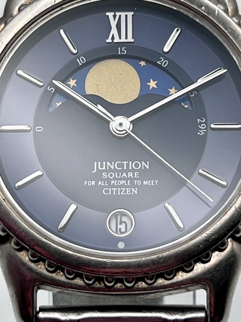 May include: Close-up of a Citizen Junction Square watch. The watch face is a gradient of dark blue to grey, featuring a moon phase indicator with a gold moon and stars. The watch has silver hands and markers, with the date displayed at the 6 o'clock position.