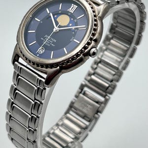 May include: A silver Citizen Junction watch with a dark blue face. The watch has a moon phase indicator and a stainless steel strap. The watch face includes the text "JUNCTION FOR ALL SQUARE PEOPLE TO MEET CITIZEN".