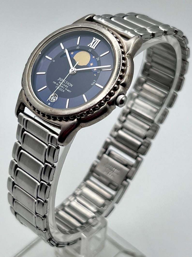 May include: A silver Citizen Junction watch with a dark blue face. The watch has a moon phase indicator and a stainless steel strap. The watch face includes the text "JUNCTION FOR ALL SQUARE PEOPLE TO MEET CITIZEN".