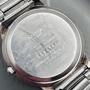 May include: A close-up of the back of a silver watch with a metal strap. The watch face is inscribed with "JUNCTION SQUARE", "WATER RESIST", and the number "3020741".