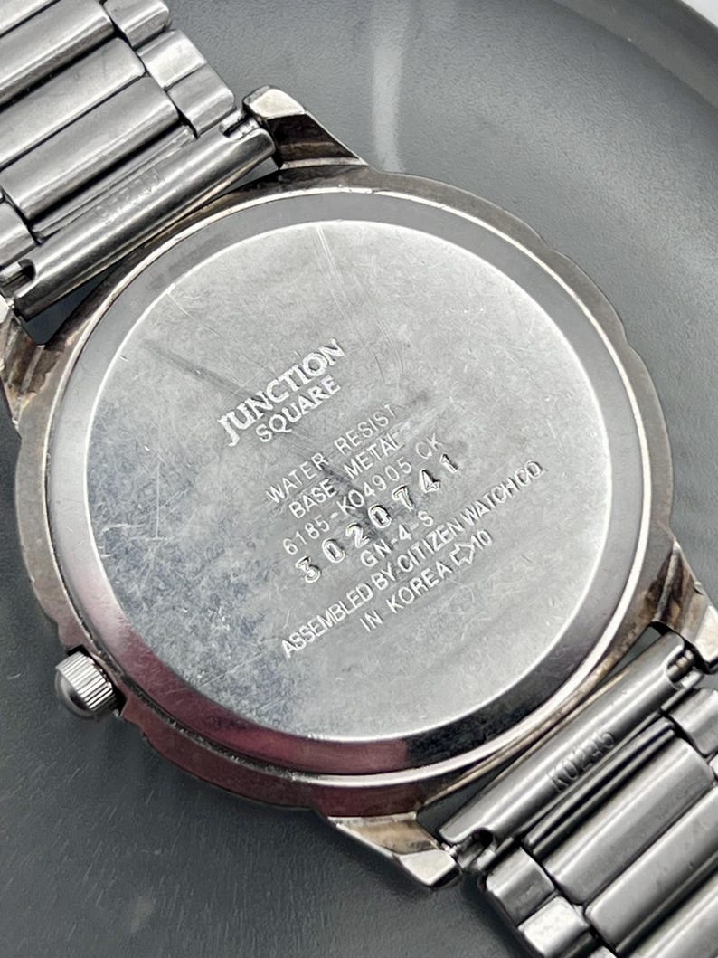 May include: A close-up of the back of a silver watch with a metal strap. The watch face is inscribed with "JUNCTION SQUARE", "WATER RESIST", and the number "3020741".