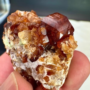 May include: A raw mineral specimen featuring a prominent, deep reddish-brown, multifaceted crystal cluster set against a base of smaller, translucent amber and white crystalline formations with a jagged, uneven texture and reflective surfaces.