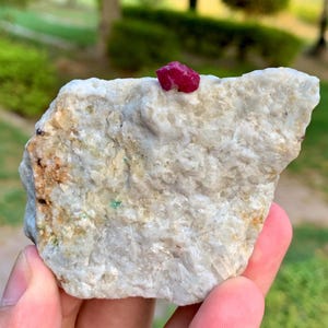 Può includere: Un esemplare minerale grezzo e non tagliato che presenta un prominente cristallo rosso magenta vibrante incastonato in una grande matrice rocciosa cristallina bianco sporco e marrone chiaro, con superfici granulari visibili e sottili inclusioni terrose.