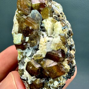 Może przedstawiać: Surowy okaz minerału z klastrami półprzezroczystych, ciemnobursztynowych i oliwkowozielonych struktur krystalicznych osadzonych w teksturowanej, jasnej matrycy. Geometryczne kryształy wykazują ostre, wielościenne krawędzie oraz błyszczące wykończenie.
