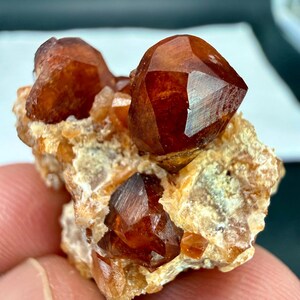 May include: A raw mineral specimen featuring deep reddish-brown, translucent, faceted-looking crystals embedded in a lighter, textured, tan-coloured matrix. The cluster displays multiple sharp, angular crystal points with a glossy, reflective surface.