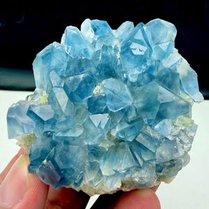 May include: A cluster of translucent, light blue crystalline structures with sharp, geometric facets. The mineral formation features varying shades of sky blue and pale azure, with a rough, pale, and textured base supporting the pointed crystals.