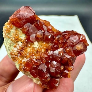 Puede incluir: Un racimo de cristales de granate de color rojo anaranjado intenso incrustados en una matriz mineral texturizada de color verde claro. El espécimen presenta múltiples puntas de cristal afiladas y facetadas con un aspecto brillante.