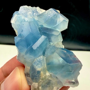 May include: A cluster of translucent, light blue fluorite crystals with sharp, geometric edges and a glassy texture. The mineral specimen features varying shades of blue, ranging from pale sky blue to deeper azure, with a white, rocky base.