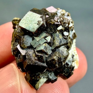 Może przedstawiać: Skupisko ciemnych oliwkowozielonych i metalicznie brązowych kryształów granatu o sześciennej strukturze. Okaz minerału posiada wiele ostrych, geometrycznych faset, które odbijają światło, ukazując głęboką, ziemistą paletę barw oraz krystaliczną teksturę.