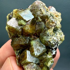 Może przedstawiać: Skupisko surowych, oliwkowozielonych kryształów granatu o półprzezroczystym, wielościennym wyglądzie. Okaz minerału charakteryzuje się ostrymi, geometrycznymi formacjami kryształów o głębokich brązowych i ziemistozielonych odcieniach, ukazując naturalną teksturę.