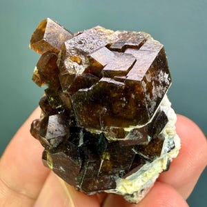 Może przedstawiać: Skupisko ciemnobrązowych, półprzezroczystych, sześciennych kryształów granatu o ostrych, geometrycznych fasetach. Okaz minerału ma błyszczącą, odblaskową powierzchnię i jest przymocowany do jasnej, szorstkiej, krystalicznej podstawy matrycowej.
