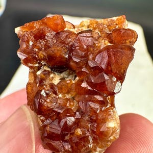 Pode incluir: Um aglomerado de cristais de granada espessartita laranja-escuro e castanho-avermelhado com uma aparência translúcida e facetada. O espécime mineral apresenta numerosas pequenas formações cristalinas angulares que crescem sobre uma base de matriz clara.
