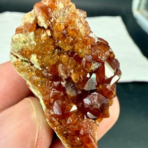 Op de afbeelding: Een ruw mineraal exemplaar met een cluster van dieporanje en roodbruine kristalstructuren. De formatie vertoont talloze kleine, gefacetteerde punten met een doorschijnende, glasachtige glans, bevestigd op een bleke, getextureerde rotsachtige basis.