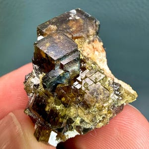 Może przedstawiać: Surowy okaz minerału z grupą sześciennych kryształów w odcieniach ciemnego brązu, oliwkowej zieleni i półprzezroczystego bursztynu. Struktura wykazuje ostre geometryczne krawędzie oraz warstwową, krystaliczną formację o naturalnej teksturze.