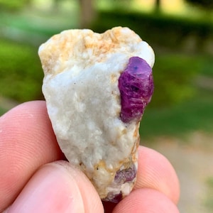 May include: A raw mineral specimen featuring a prominent, deep magenta-coloured crystal embedded within a textured, opaque white and beige matrix. The stone has an irregular, jagged shape with a rough surface texture and visible crystalline inclusions.