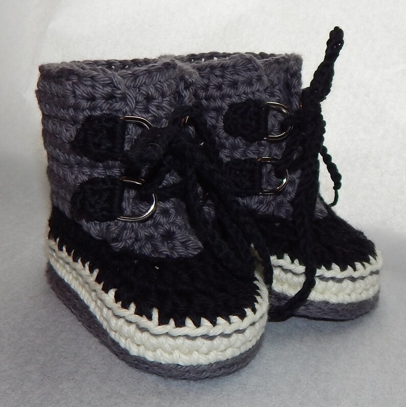 K&ouml;nnte beinhalten: Ein Paar graue und schwarze geh&auml;kelte Babyschuhe mit wei&szlig;em Besatz. Die Schuhe haben ein Schn&uuml;rdesign mit silbernen Schnallen.