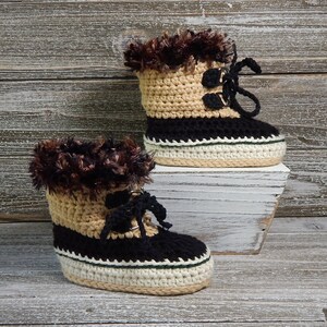 K&ouml;nnte beinhalten: Ein Paar geh&auml;kelte Babyschuhe mit braunem Fellbesatz, schwarzen und braunen Streifen und einem wei&szlig;en und gr&uuml;nen Besatz um die Sohle.