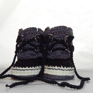 K&ouml;nnte beinhalten: Ein Paar graue und schwarze geh&auml;kelte Babyschuhe mit wei&szlig;em Besatz und schwarzen Schn&uuml;rsenkeln. Die Schuhe haben silberne Metall&ouml;sen und sind mit einer schwarzen Schnur zusammengebunden.