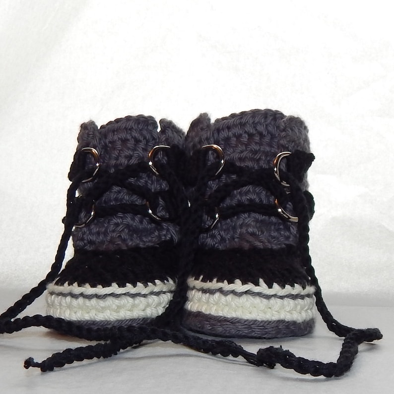 K&ouml;nnte beinhalten: Ein Paar graue und schwarze geh&auml;kelte Babyschuhe mit wei&szlig;em Besatz und schwarzen Schn&uuml;rsenkeln. Die Schuhe haben silberne Metall&ouml;sen und sind mit einer schwarzen Schnur zusammengebunden.