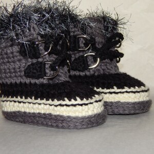 K&ouml;nnte beinhalten: Ein Paar graue geh&auml;kelte Babyschuhe mit schwarzem Besatz und wei&szlig;en Akzenten. Die Schuhe haben silberne Schnallen und schwarze Schn&uuml;rsenkel. Die Schuhe sind mit einem flauschigen grauen und schwarzen Besatz verziert.