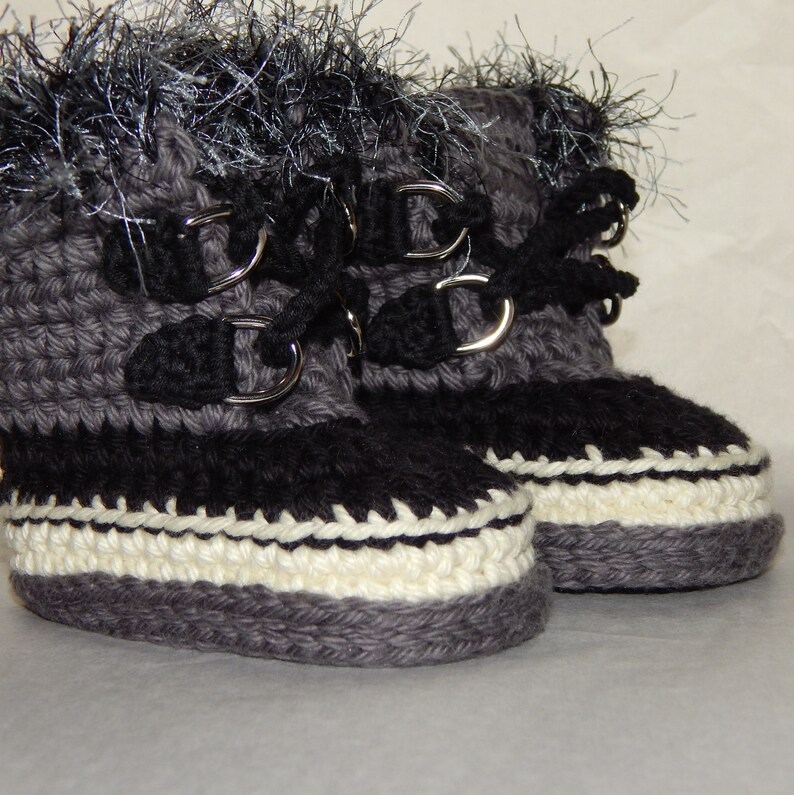 K&ouml;nnte beinhalten: Ein Paar graue geh&auml;kelte Babyschuhe mit schwarzem Besatz und wei&szlig;en Akzenten. Die Schuhe haben silberne Schnallen und schwarze Schn&uuml;rsenkel. Die Schuhe sind mit einem flauschigen grauen und schwarzen Besatz verziert.