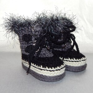 K&ouml;nnte beinhalten: Ein Paar graue und schwarze geh&auml;kelte Babyschuhe mit wei&szlig;en R&auml;ndern und schwarzen Schn&uuml;rsenkeln. Die Schuhe haben einen flauschigen wei&szlig;en und grauen Besatz am oberen Rand.