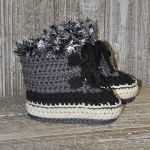 K&ouml;nnte beinhalten: Graue, schwarze und wei&szlig;e geh&auml;kelte Babyschuhe mit einem flauschigen Besatz am oberen Rand. Die Schuhe haben Schn&uuml;rsenkel und eine wei&szlig;e Sohle.