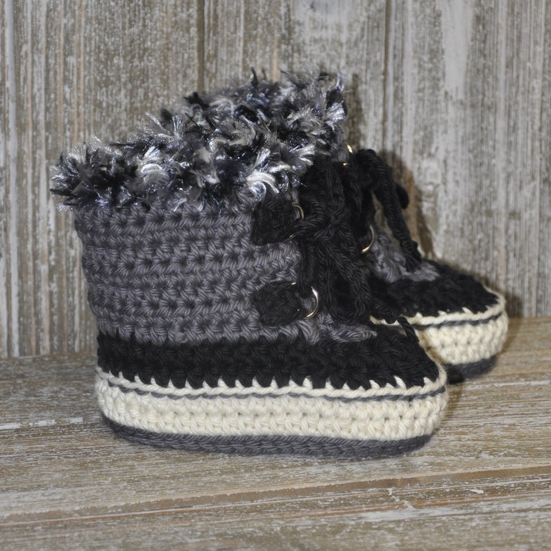 K&ouml;nnte beinhalten: Graue, schwarze und wei&szlig;e geh&auml;kelte Babyschuhe mit einem flauschigen Besatz am oberen Rand. Die Schuhe haben Schn&uuml;rsenkel und eine wei&szlig;e Sohle.