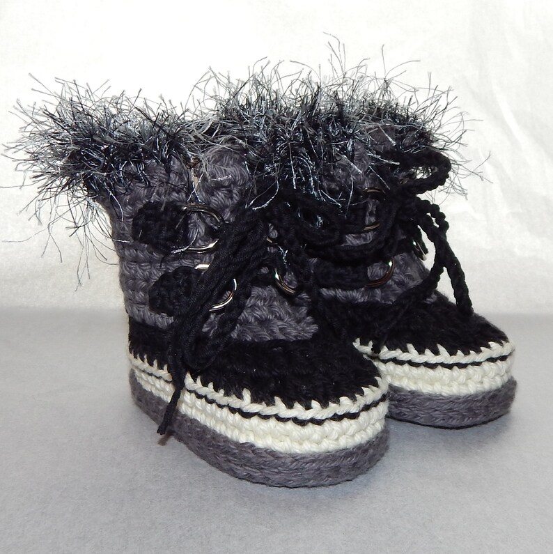 K&ouml;nnte beinhalten: Ein Paar graue und schwarze geh&auml;kelte Babyschuhe mit wei&szlig;em Besatz und schwarzen Schn&uuml;rsenkeln. Die Schuhe haben einen flauschigen wei&szlig;en und grauen Besatz am oberen Rand.