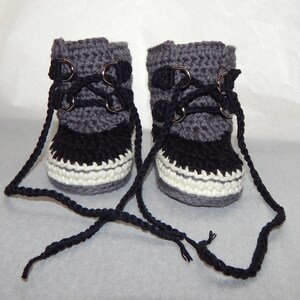 K&ouml;nnte beinhalten: Ein Paar graue und schwarze geh&auml;kelte Babyschuhe mit wei&szlig;em Besatz und schwarzen Schn&uuml;rsenkeln. Die Schuhe haben Metall&ouml;sen und sind mit den Schn&uuml;rsenkeln zusammengebunden.