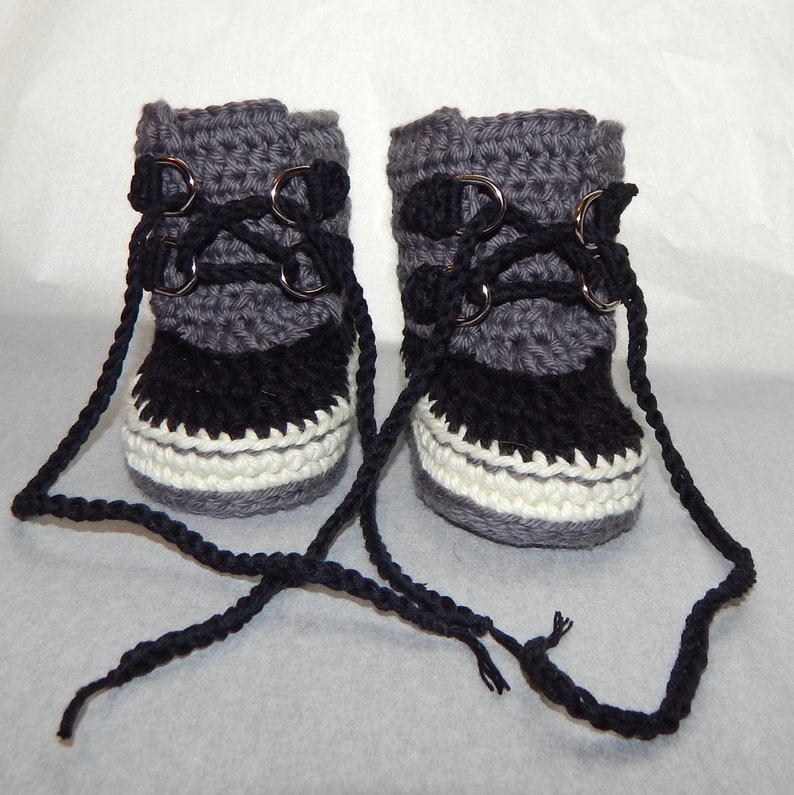 K&ouml;nnte beinhalten: Ein Paar graue und schwarze geh&auml;kelte Babyschuhe mit wei&szlig;em Besatz und schwarzen Schn&uuml;rsenkeln. Die Schuhe haben Metall&ouml;sen und sind mit den Schn&uuml;rsenkeln zusammengebunden.