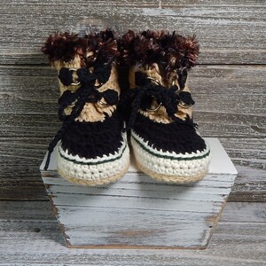 K&ouml;nnte beinhalten: Ein Paar geh&auml;kelte Babyschuhe mit braunem Fellbesatz, schwarzem und braunem Garn und wei&szlig;en und gr&uuml;nen Akzenten.