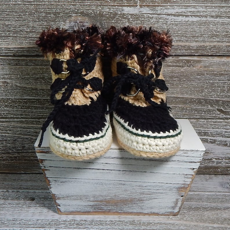 K&ouml;nnte beinhalten: Ein Paar geh&auml;kelte Babyschuhe mit braunem Fellbesatz, schwarzem und braunem Garn und wei&szlig;en und gr&uuml;nen Akzenten.