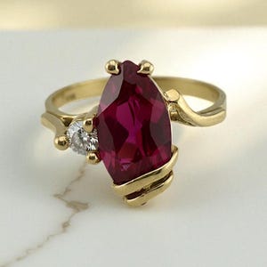 Puede incluir: Un anillo de oro con una gran piedra preciosa roja oscura con corte marquesa. Una piedra preciosa más pequeña, redonda y transparente está engastada a un lado. El anillo tiene una banda de oro retorcida y detalles dorados decorativos.