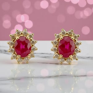 Puede incluir: Un par de pendientes de botón dorados. Cada pendiente presenta una vibrante piedra roja de forma ovalada rodeada por un halo de piedras más pequeñas y transparentes. Los pendientes están sobre un fondo rosa suave.