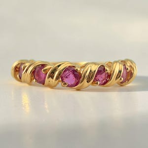 May include: A gold ring with a twisted design, featuring several round, bright pink gemstones. The ring's band has a textured, swirling pattern, and the gemstones are evenly spaced along the top.