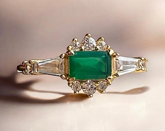 May include: A gold ring with a green emerald gemstone surrounded by a halo of smaller diamonds. The ring has a baguette diamond band.