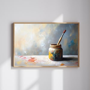 May include: A painting depicting a paintbrush resting in a jar of paint. The jar is filled with various colours of paint, and the brush is covered in paint. The background is a light blue and white, with hints of yellow and orange. The painting is framed in a light wood frame.