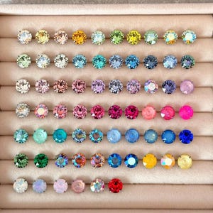 Puede incluir: Una colección de pendientes de botón coloridos dispuestos en una vitrina. Los pendientes presentan una variedad de colores, incluyendo amarillo, rosa, azul, verde y morado. Cada pendiente tiene una gema redonda y facetada.