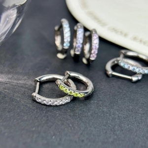 May include: Silver hoop earrings with various colored gemstones. One pair features white stones, another has green stones, and others display light blue and purple gems. The earrings are displayed on a dark surface.