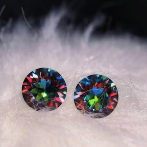 May include: Two round gemstones displaying a spectrum of colours, including blue, green, red, and yellow. The gems are set against a soft, white background, creating a striking contrast. The gemstones appear to be faceted, reflecting light.