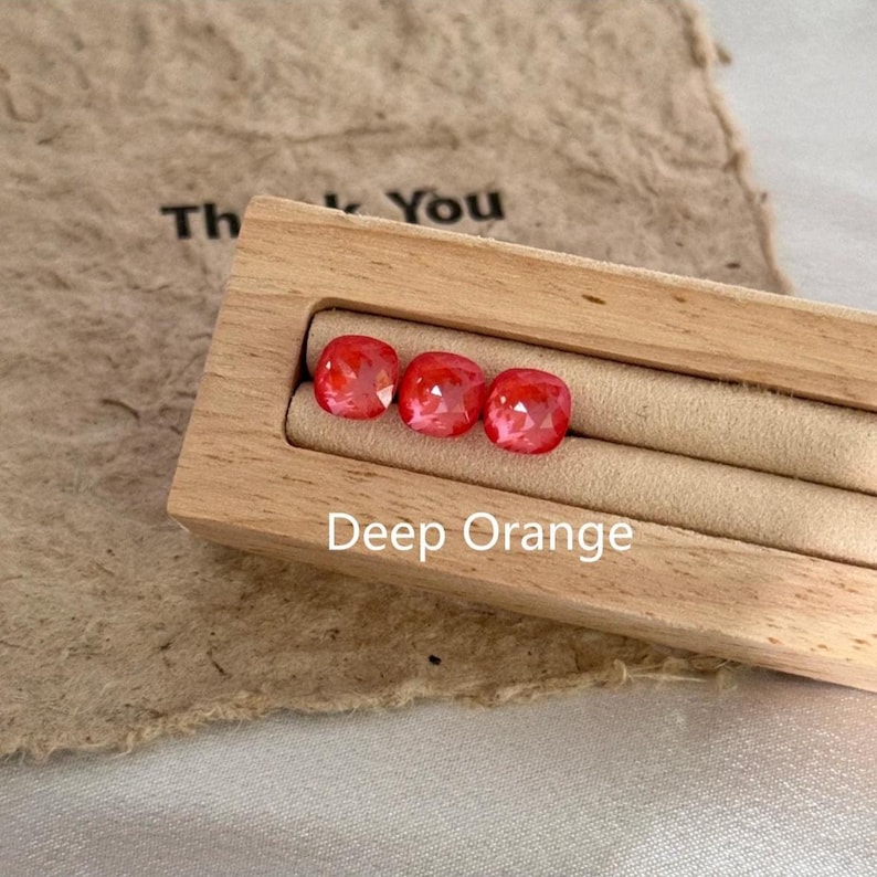 May include: Three deep orange gemstones with a faceted cut are displayed in a wooden box. The box sits on a textured, tan surface. The text "Deep Orange" is visible below the gemstones.
