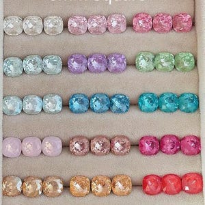 May include: Rows of 8mm square gemstones in various colors, including clear, pink, purple, green, blue, and orange. The gemstones are displayed in a beige tray, showcasing their faceted surfaces and vibrant hues.