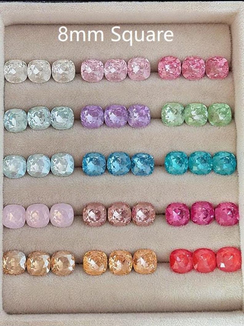 May include: Rows of 8mm square gemstones in various colors, including clear, pink, purple, green, blue, and orange. The gemstones are displayed in a beige tray, showcasing their faceted surfaces and vibrant hues.