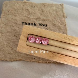 May include: Three light pink gemstones are displayed in a wooden holder. The holder is labeled "Light Pink" and is set against a textured paper background with the words "Thank You" printed on it. The gemstones are square-cut.