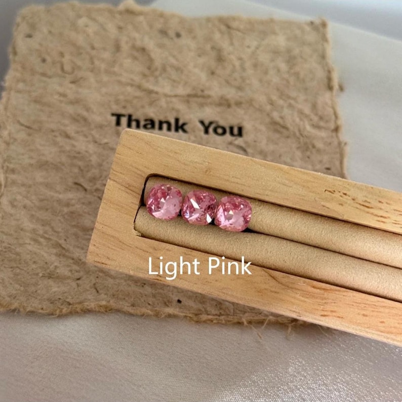 May include: Three light pink gemstones are displayed in a wooden holder. The holder is labeled "Light Pink" and is set against a textured paper background with the words "Thank You" printed on it. The gemstones are square-cut.