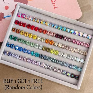 May include: A rectangular grey velvet display tray containing seven horizontal rows of round, faceted, multi-coloured rhinestones. The stones are arranged in a gradient of colours including red, orange, yellow, green, blue, and purple. Text reads: BUY 1 GET 1 FREE (Random Colors).